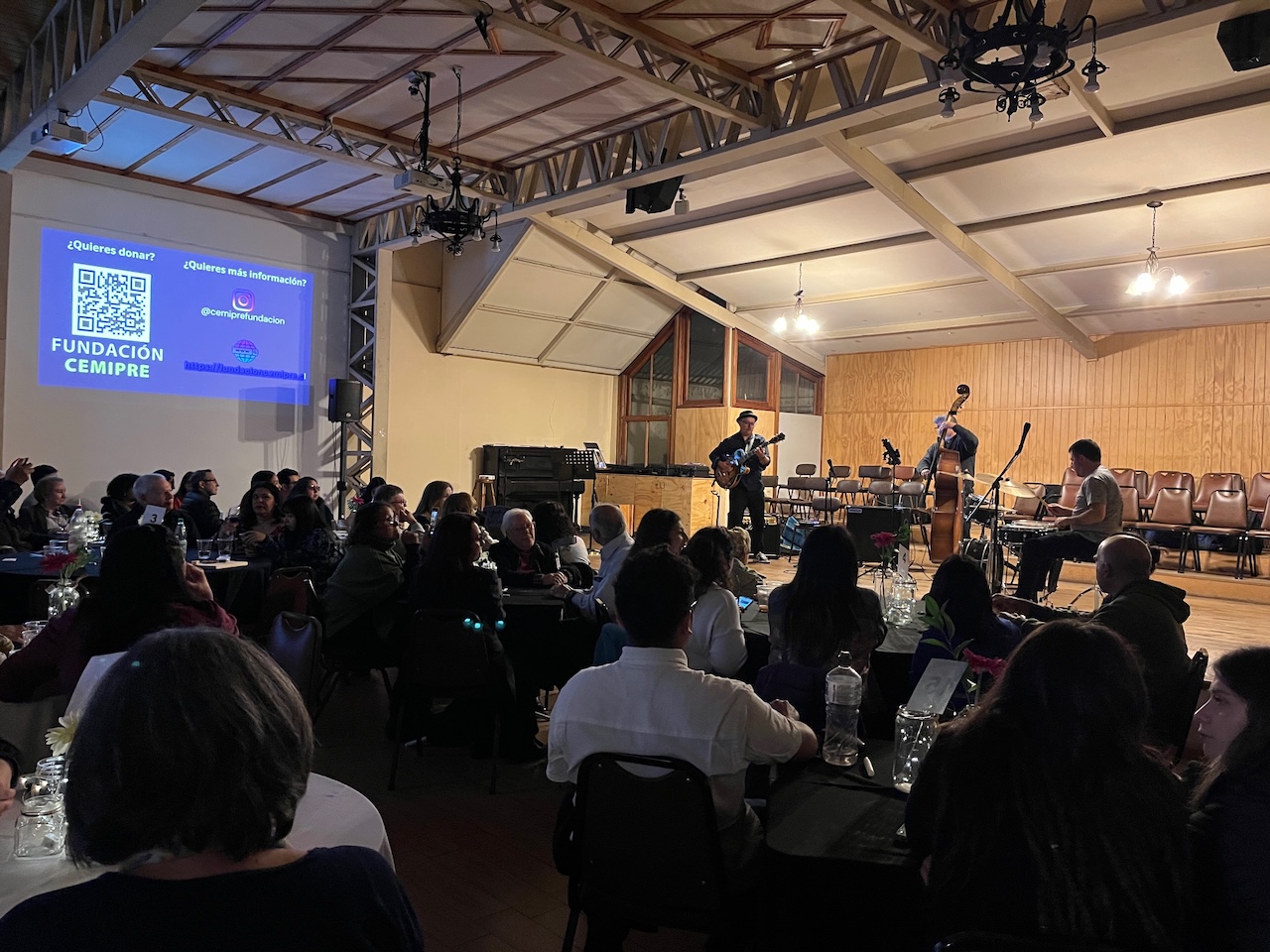Vista del salón desde atras, 80 personas sentadas en mesas, musicos en el escenario tocando.